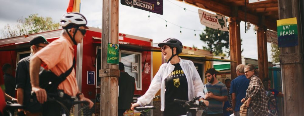 A photo os Sarah standing with her bike and helmet at the Portland Marcado. She is wearing a "Sunrise Movement PDX" shirt and smiling at another cyclist who is wearing his helmet.
