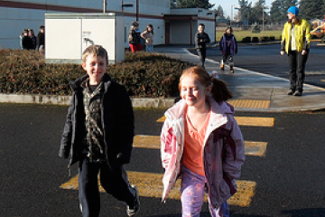 Children are seen walking across a crosswalk on a fall day wearing their heavier coats and smiling as the sun shines on them.
