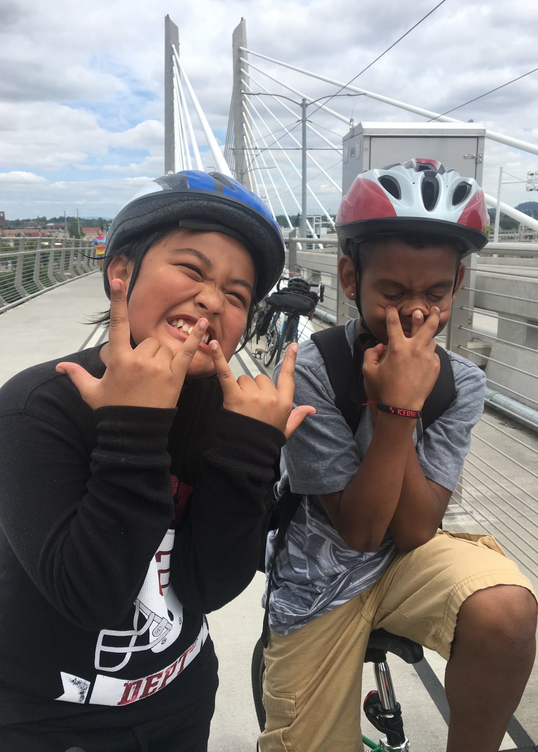 Two children stand on the Tillikum Bridge. They both wear bike helmets. One child is making an enthusiastic smile, squinting their eyes and holding up rock and roll hands.