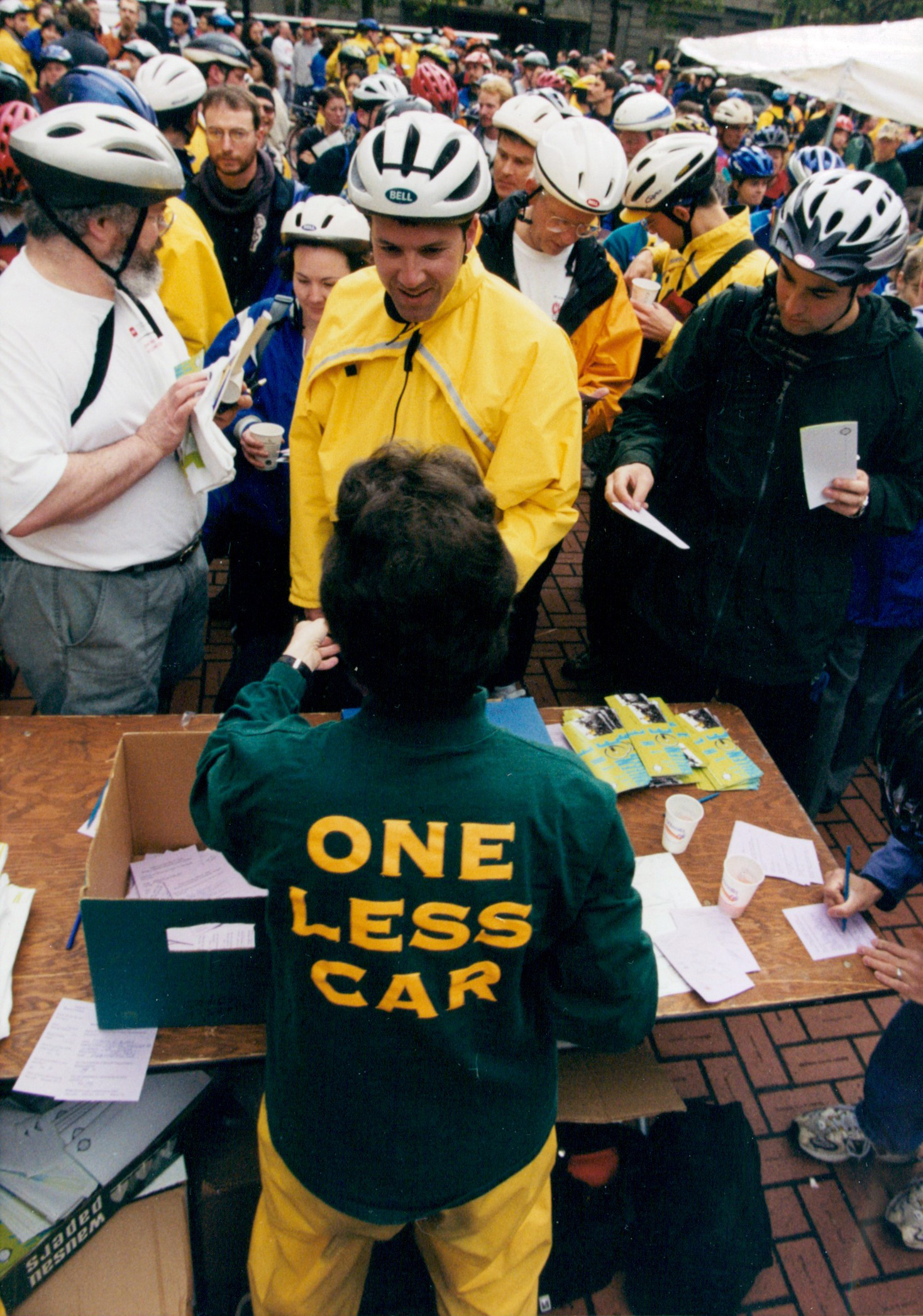 A large group of many people wearing bike helmets. There are so many it is hard to distinguish between them. They are facing a table with a variety of literature on it, but none of the titles or words on the literature are visible. One person stands on the near side of the table, facing all the bicyclists. The person is wearing a jacket that reads "One Less Car"