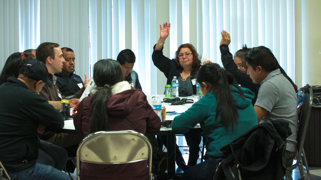 A diverse group of people sit around a table, seated in folding chairs. Two people have their hands raised. Others look at them, look at the table, or look away.